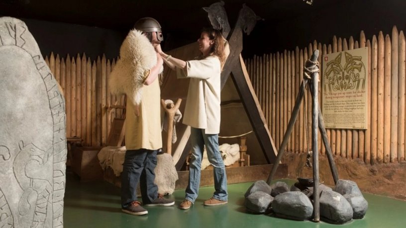 Women helping man dressing up as Viking warrior next to a fireplace setup for cooking 