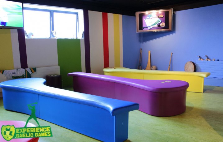 Colourful Room for Group of Players - Experience Gaelic Games Blue Yellow and Violet Curvy Benches and Stripy walls with a  TV/Monitor on the Wall - Green Floor